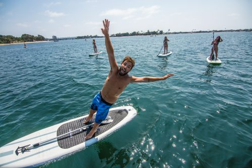 Man facing the camera diving off a paddle board into the sea, with other people on paddle boards in the background